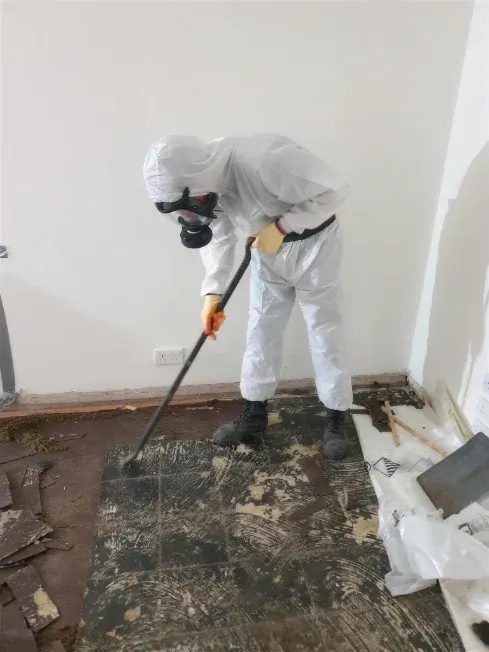 Operative in full coveralls and respirator lifting bitumen-backed floor tiles from a room