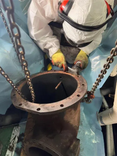 Operative in protective coveralls removing an asbestos gasket from a flanged pipe in a ship&rsquo;s engine room, working over protective sheeting